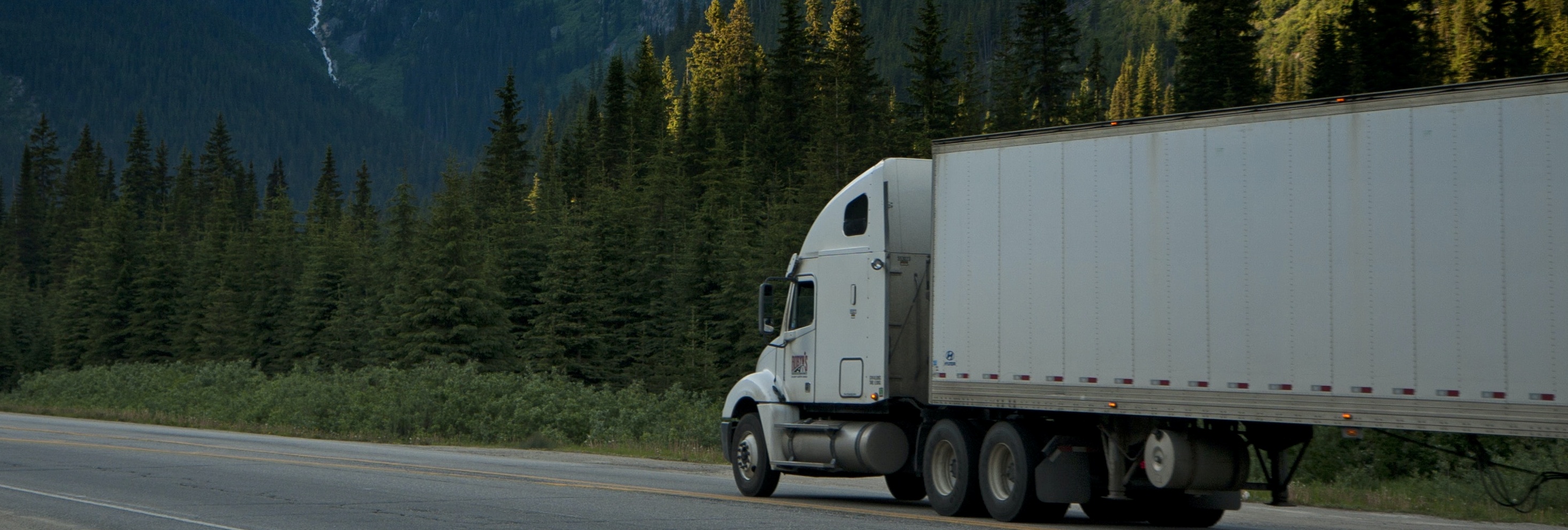 A semi-truck driving along a winding road lined with lush green trees and mountains in the background.